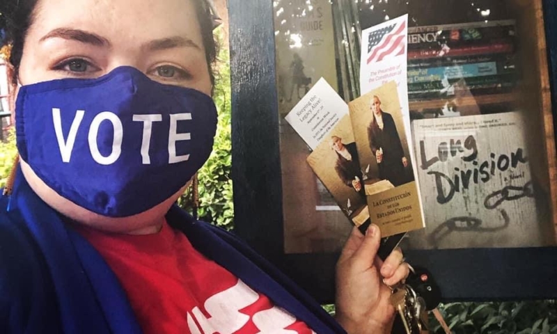 Woman wearing a "VOTE" mask holding up pocket constitutions in front of a Little Free Library